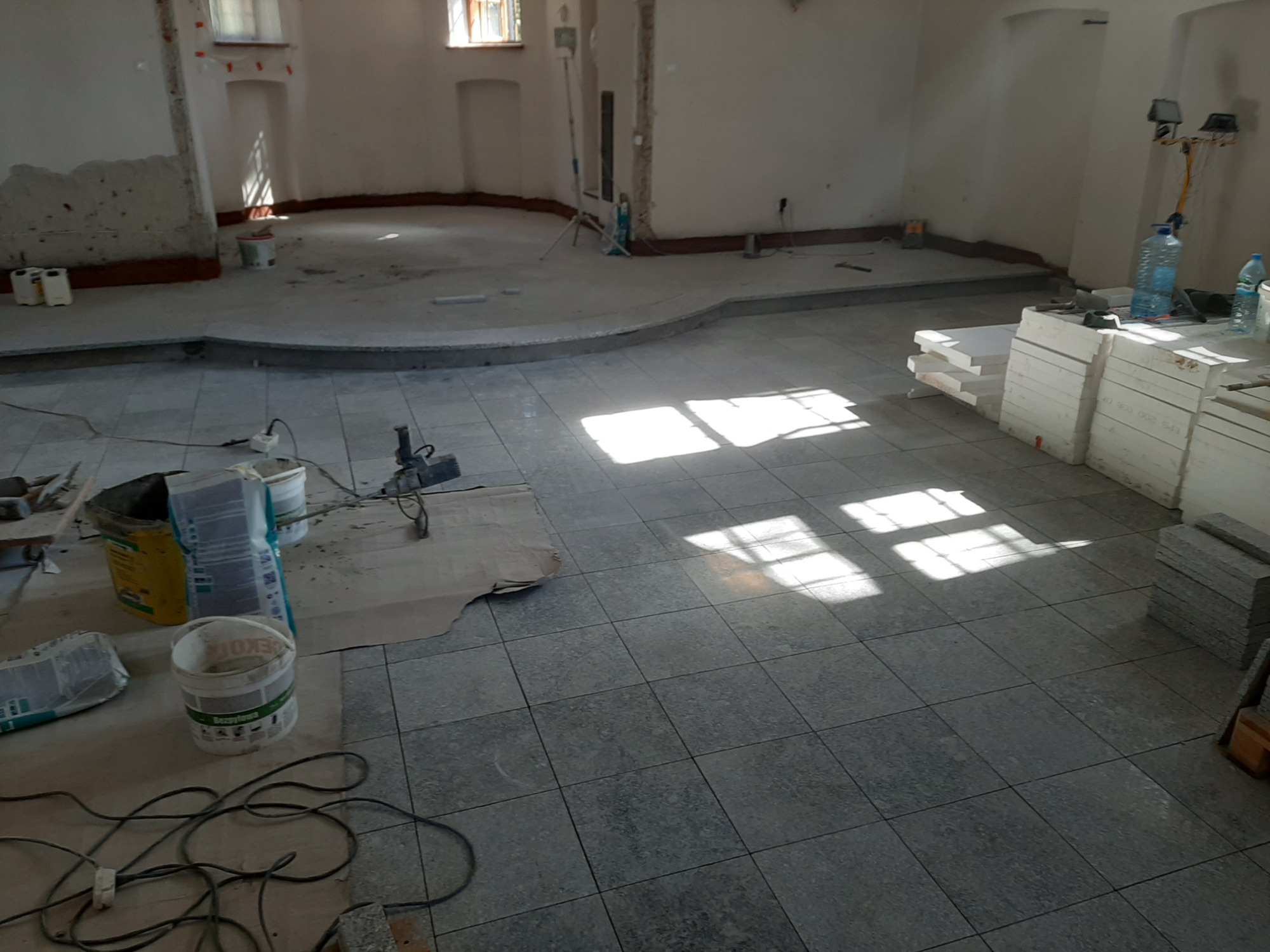 Wnętrze w trakcie prac wykończeniowych, szare płytki na podłodze, narzędzia i materiały budowlane w nieładzie, widoczne promienie słoneczne padające przez okno.