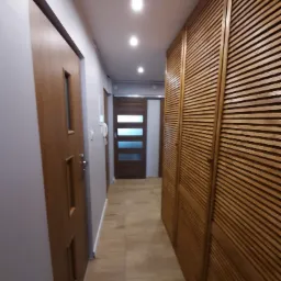 Wąski korytarz z drewnianą podłogą, po lewej stronie drzwi z małymi okienkami, po prawej zabudowa szafy z lamelami, w tle widoczne drzwi z przeszkleniami.