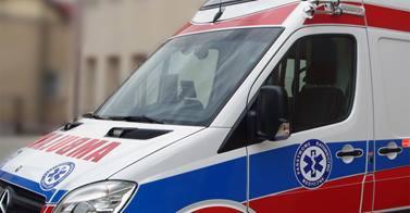 Ambulans z widocznym symbolem medycznym na boku i z przodu, zaparkowany przed budynkiem. Ujęcie z bliska, skupiające się na przedniej części pojazdu.