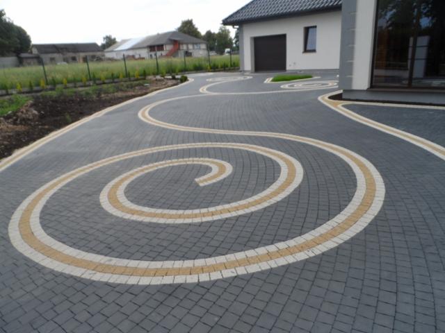 Podjazd z szarej kostki brukowej ułożonej w spiralny wzór z elementami jasnej kostki, prowadzący do nowoczesnego domu z garażem.
