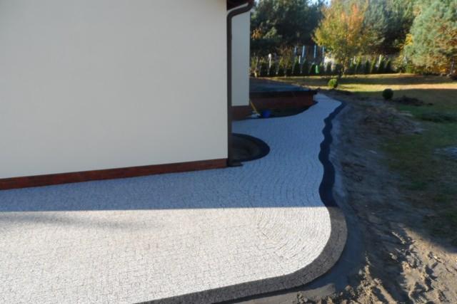 Ułożona jasnoszara kostka brukowa tworząca ścieżkę wokół domu z ciemnoszarym obramowaniem, widok z boku budynku i fragment trawnika.