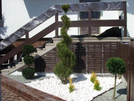 Aranżacja ogrodu z białym żwirem, formowanymi krzewami i schodami z drewnianą balustradą zabezpieczoną folią.