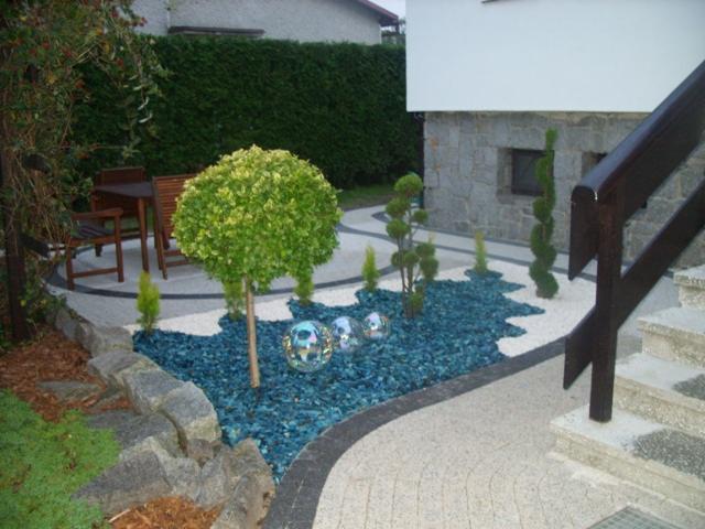 Aranżacja ogrodu z kulistym drzewkiem, niebieskim grysem i szklanymi kulami dekoracyjnymi, widoczne kamienne obrzeża i ścieżki z jasnego żwiru.