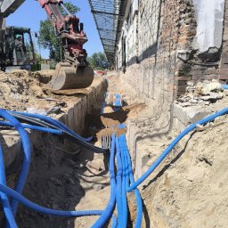 RKW Ryszard Kuba - Wykop z ułożonymi niebieskimi rurami kablowymi w ziemi, obok budynku z cegły i koparka z przodu. Prace ziemne i instalacyjne w toku.