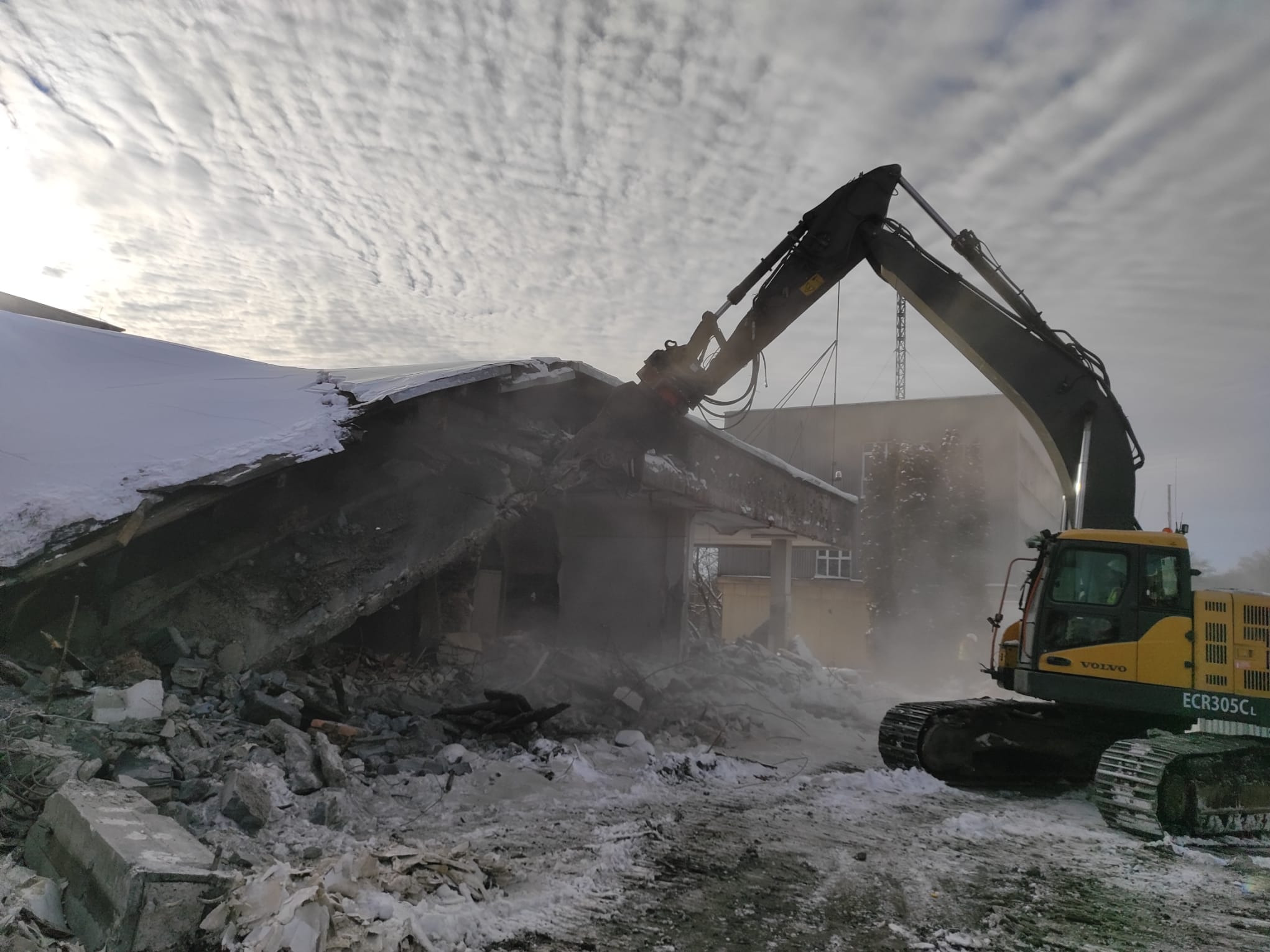 Żółta koparka Volvo ECR305CL w trakcie rozbiórki budynku, z gruzem i śniegiem w tle, pod pochmurnym niebem. Widoczny fragment sąsiedniego budynku.