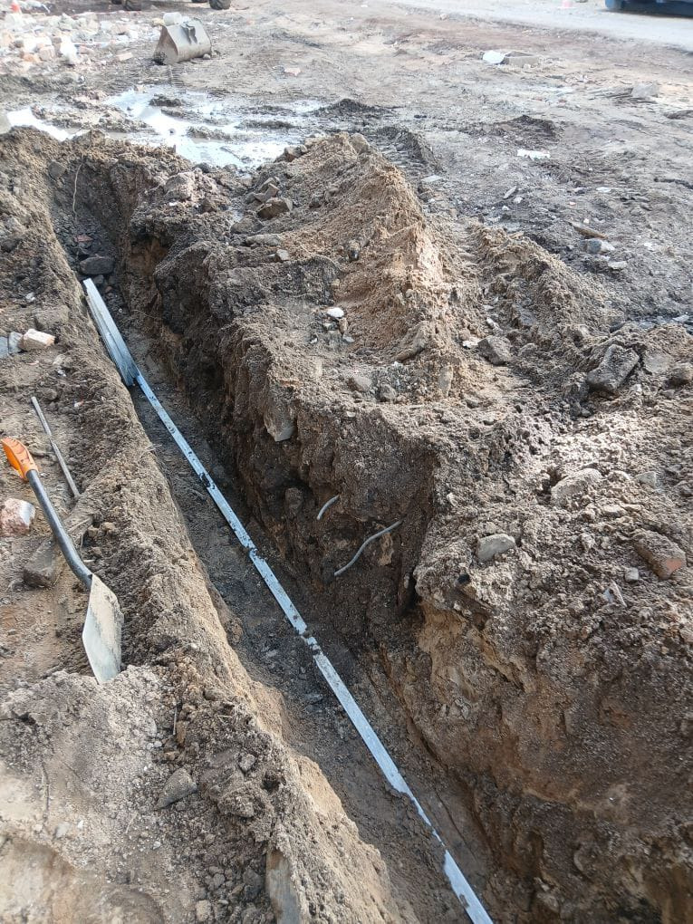 Wykop w ziemi z ułożoną rurą i kablem, widoczne narzędzia (szpadel). Ślady opon na ziemi w tle. Przygotowanie do instalacji podziemnej.