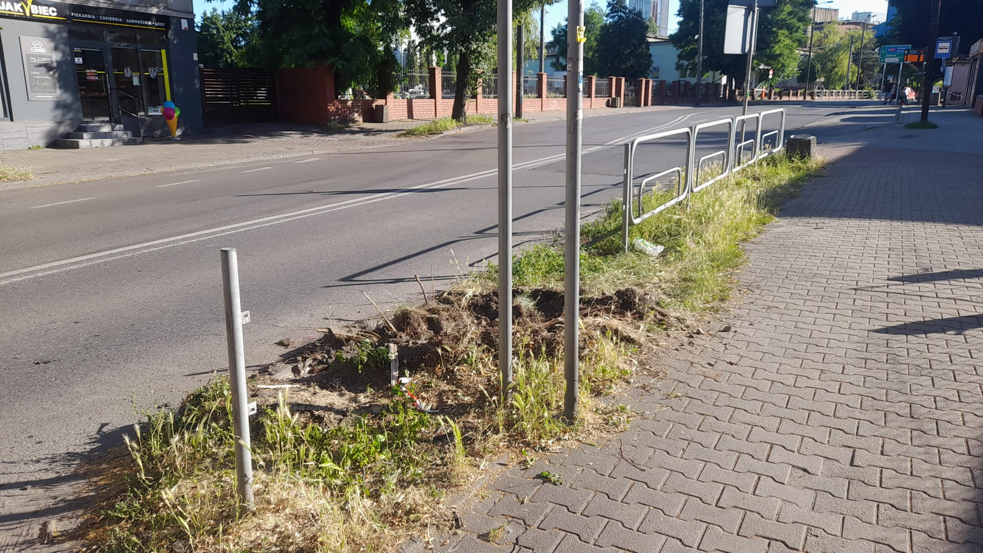 Słupki drogowe w trakcie montażu przy chodniku z kostki, widoczne wykopy ziemi i elementy mocujące. Tło stanowi ulica i budynki w oddali, w słoneczny dzień.