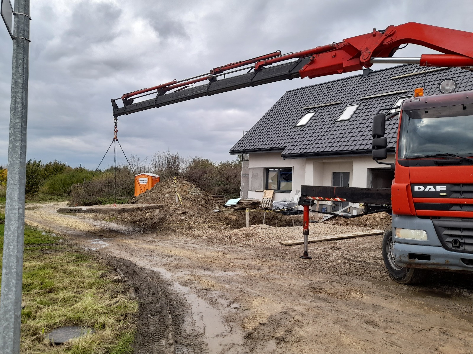 Czerwony dźwig samochodowy unosi betonowy element na placu budowy domu jednorodzinnego. W tle widoczny teren z błotem i budynek w trakcie prac wykończeniowych.