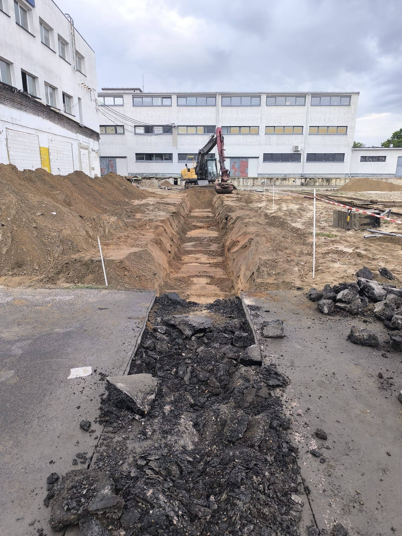 Wykop liniowy pod instalacje na terenie przemysłowym, z widocznym gruzem i koparką w tle. Prace ziemne w toku, teren zabezpieczony taśmą.