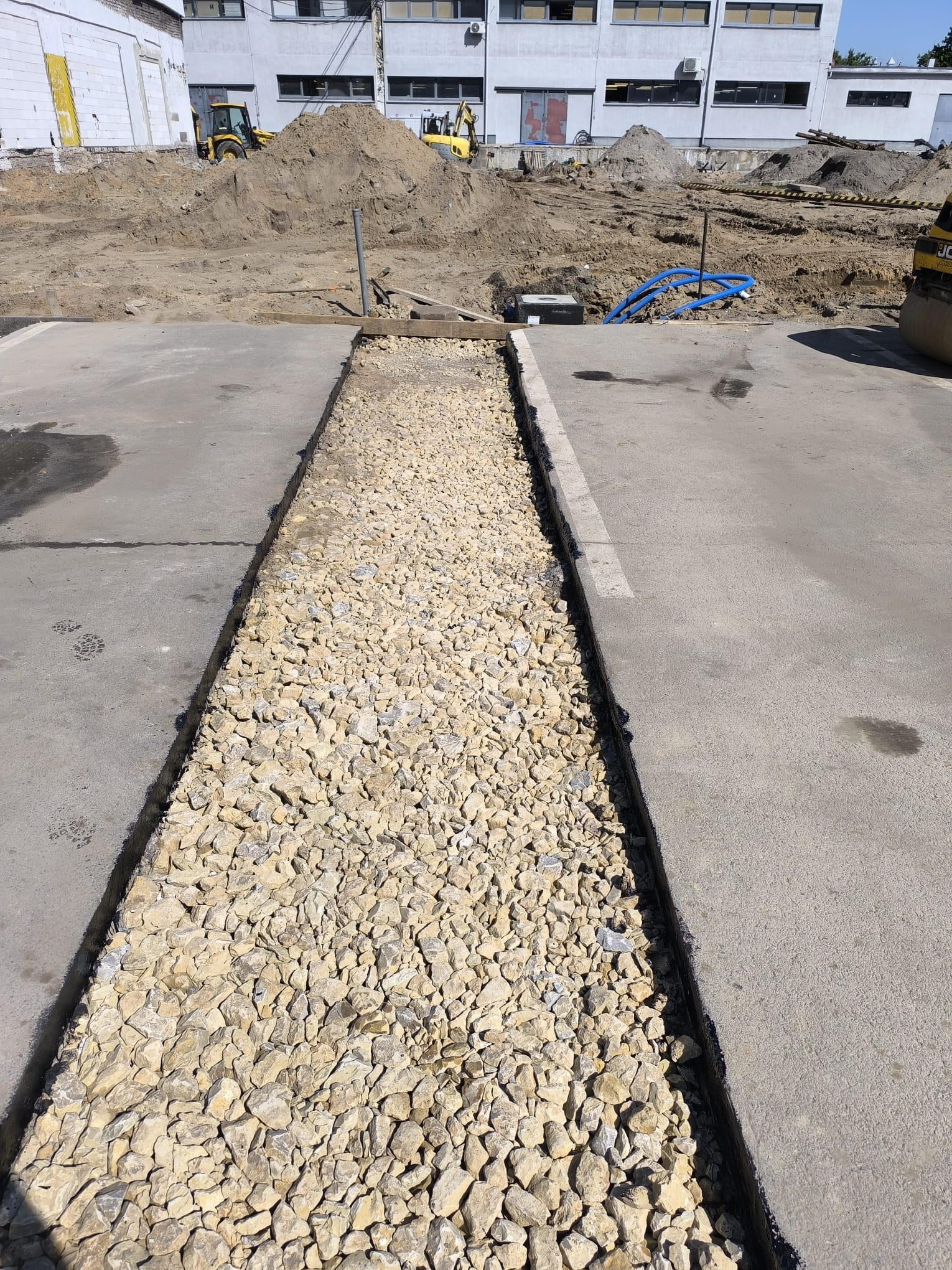 Wykop wypełniony żwirem na betonowym placu budowy, w tle maszyny budowlane i budynek. Widok z góry, oświetlenie dzienne.