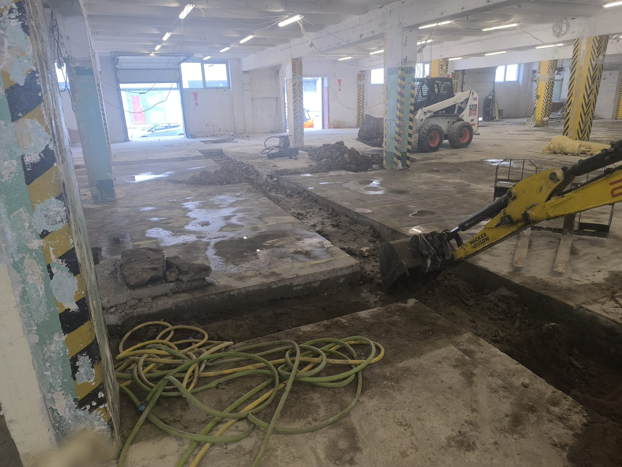 Wykop pod instalacje w hali: żółty wąż, wykopane rowy w betonie, mini koparka Wacker Neuson i Bobcat S220 w tle. Prace ziemne wewnątrz budynku.