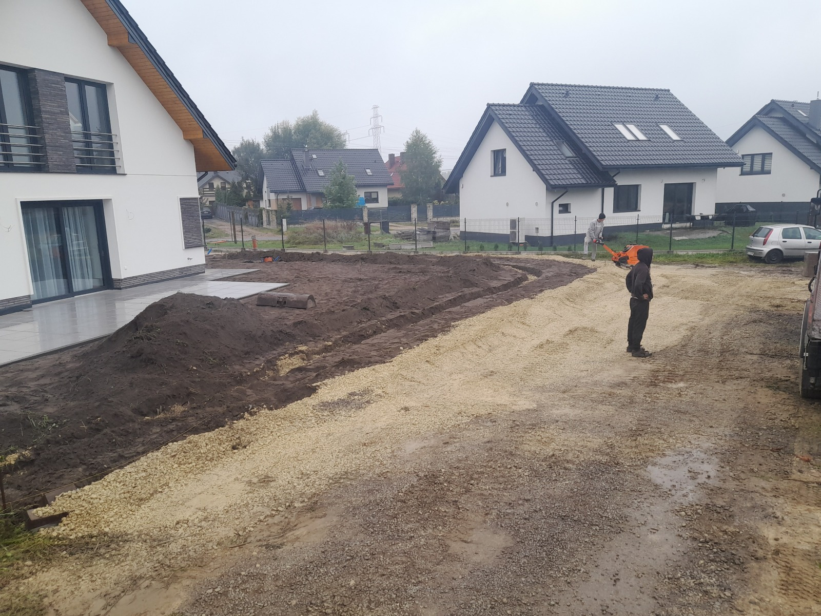 Przygotowanie podłoża pod drogę dojazdową: wyrównany teren z warstwą kruszywa, widoczne domy w tle, pracownik z zagęszczarką. Etap utwardzania drogi.