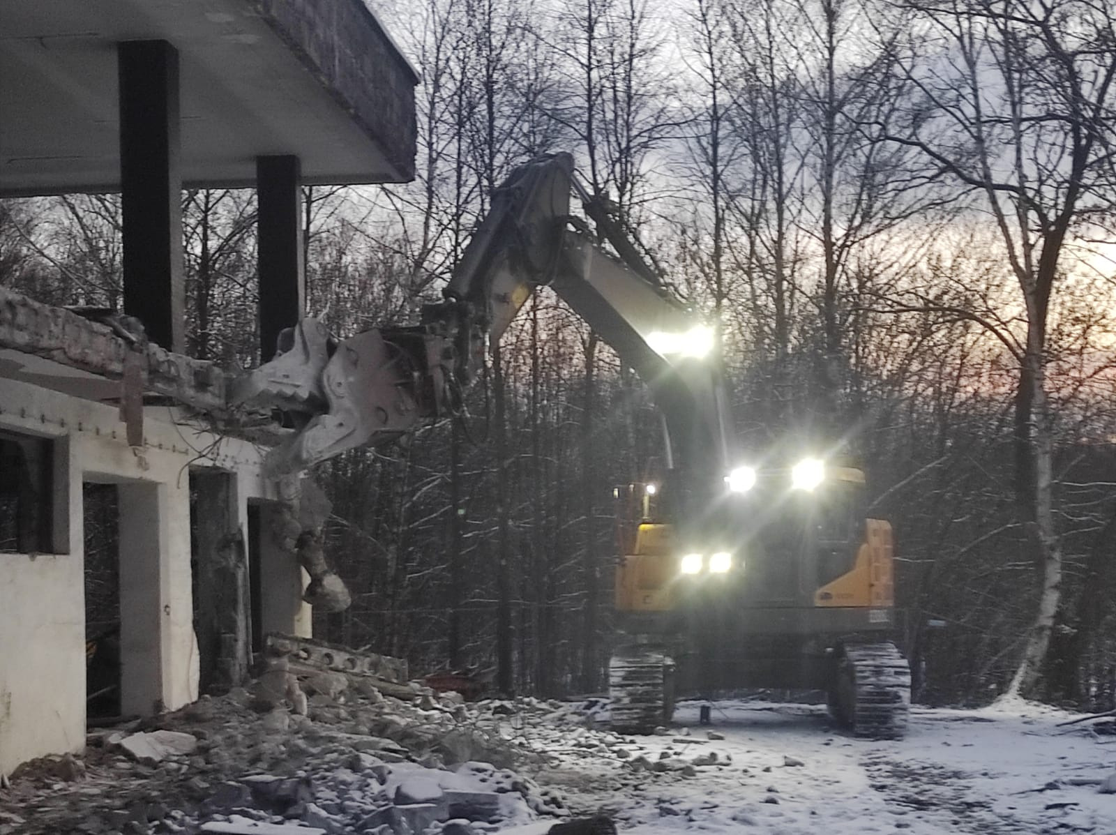 Żółta koparka z reflektorami burzy betonowy budynek w zimowej scenerii. Gruz leży na śniegu, a w tle widać drzewa.