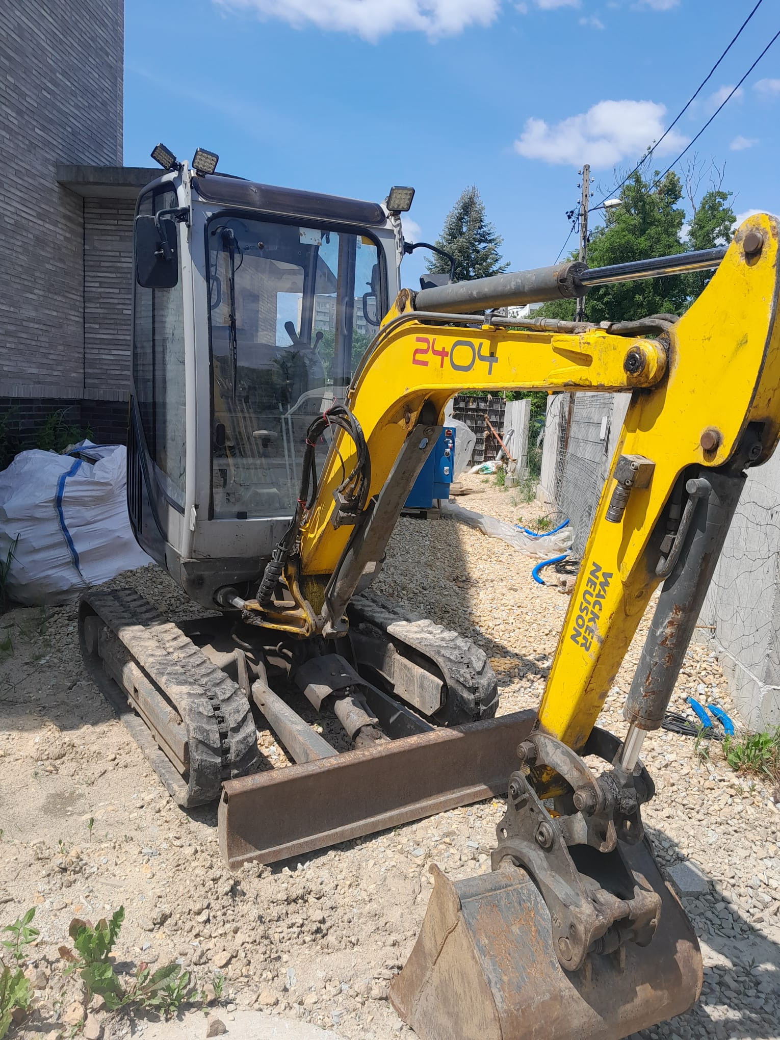Żółta minikoparka Wacker Neuson na placu budowy, widok z boku, z numerem 2404 na ramieniu, w tle materiały budowlane i fragment budynku.
