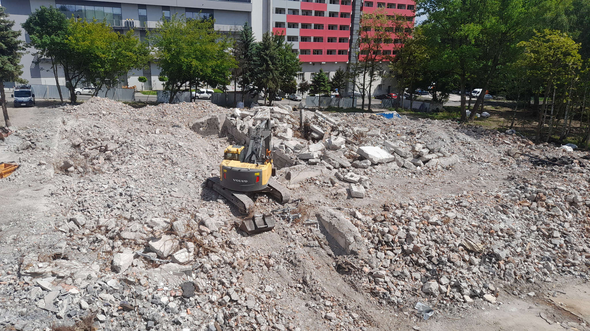 Żółta koparka Volvo na gruzowisku po wyburzeniu, z budynkami w tle. Widok z góry na plac budowy w słoneczny dzień.