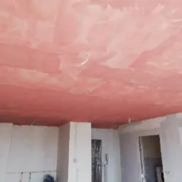Nier&oacute;wnomiernie nałożony, r&oacute;żowy tynk na suficie podczas prac remontowych w pomieszczeniu z surowymi ścianami i widocznymi przewodami.