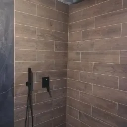 Wykończona kabina prysznicowa z brązowymi płytkami imitującymi drewno, czarną deszczownicą i baterią podtynkową, widoczne niedociągnięcia na suficie.