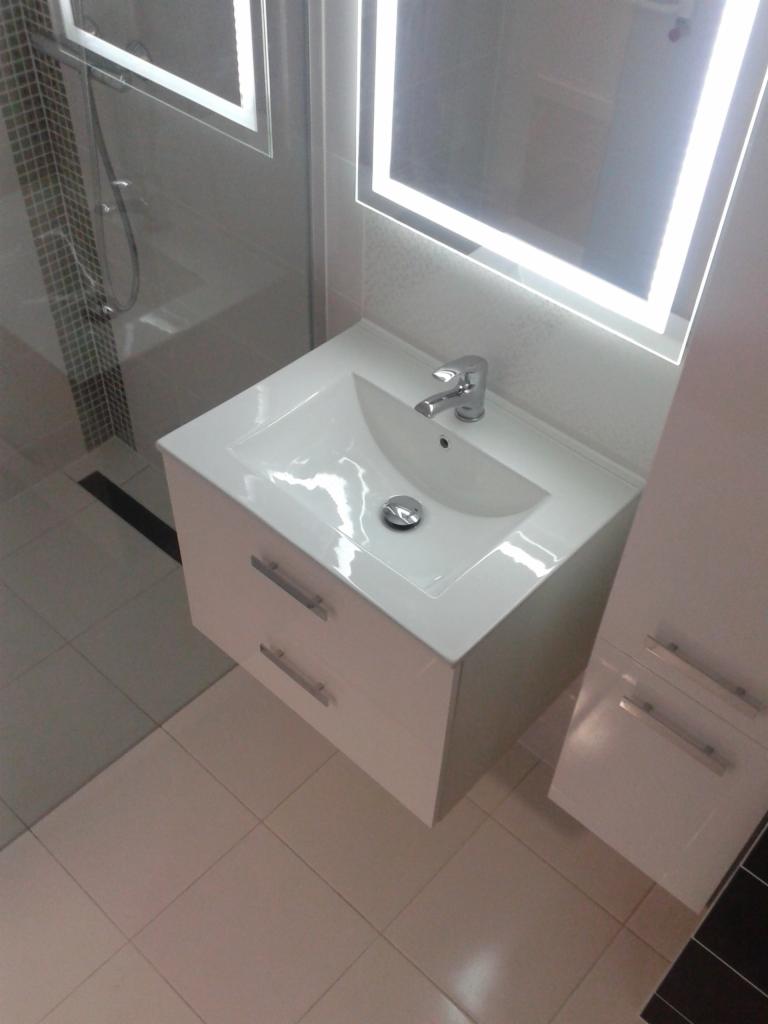 Biała umywalka z szafką i dwoma szufladami, chromowana bateria, podświetlane lustro w łazience z jasnymi płytkami i kabiną prysznicową.