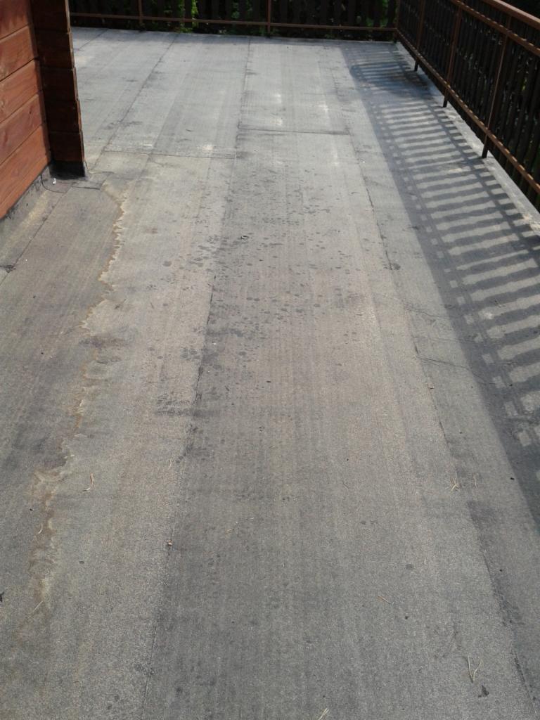 Szara, płaska powierzchnia dachu lub tarasu z widocznymi śladami wilgoci i łączeniami papy, otoczona brązową balustradą i drewnianą ścianą budynku.