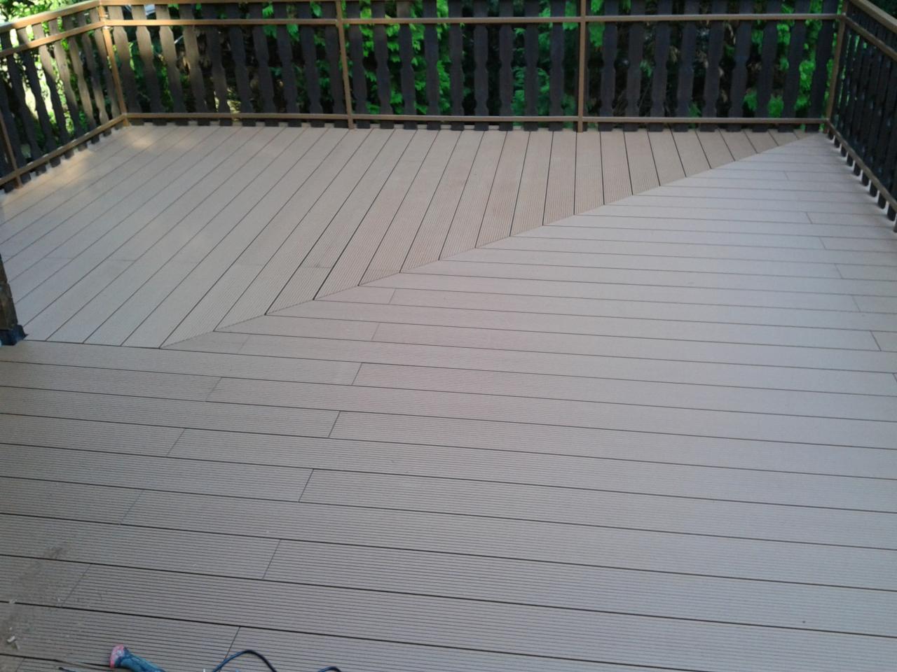 Nowoczesny taras z desek kompozytowych ułożonych w geometryczny wzór, widoczne balustrady i zieleń w tle.