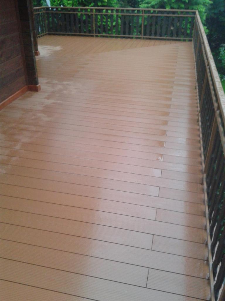 Drewniany taras z brązową balustradą po deszczu, widoczne ślady wilgoci na deskach, zieleń w tle.