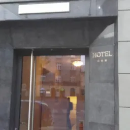 Wejście do hotelu z kamienną fasadą i szklanymi drzwiami, na których widać odbicie ulicy. Nad wejściem szyld. Po prawej stronie napis 'HOTEL' i trzy gwiazdki.