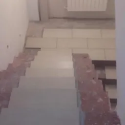 Wnętrze w trakcie wykańczania: schody z jasnymi płytkami, fragment podłogi z ułożonymi kaflami, ściany przygotowane do malowania. Widok z góry.