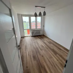 Pusty pokój po remoncie z drewnianą podłogą, białymi ścianami i trzema lampami wiszącymi, widok zza otwartych białych drzwi.