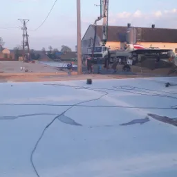 Wylewanie betonu przemysłowego z pompy na placu budowy, widoczni pracownicy, w tle budynek gospodarczy i słupy elektryczne.