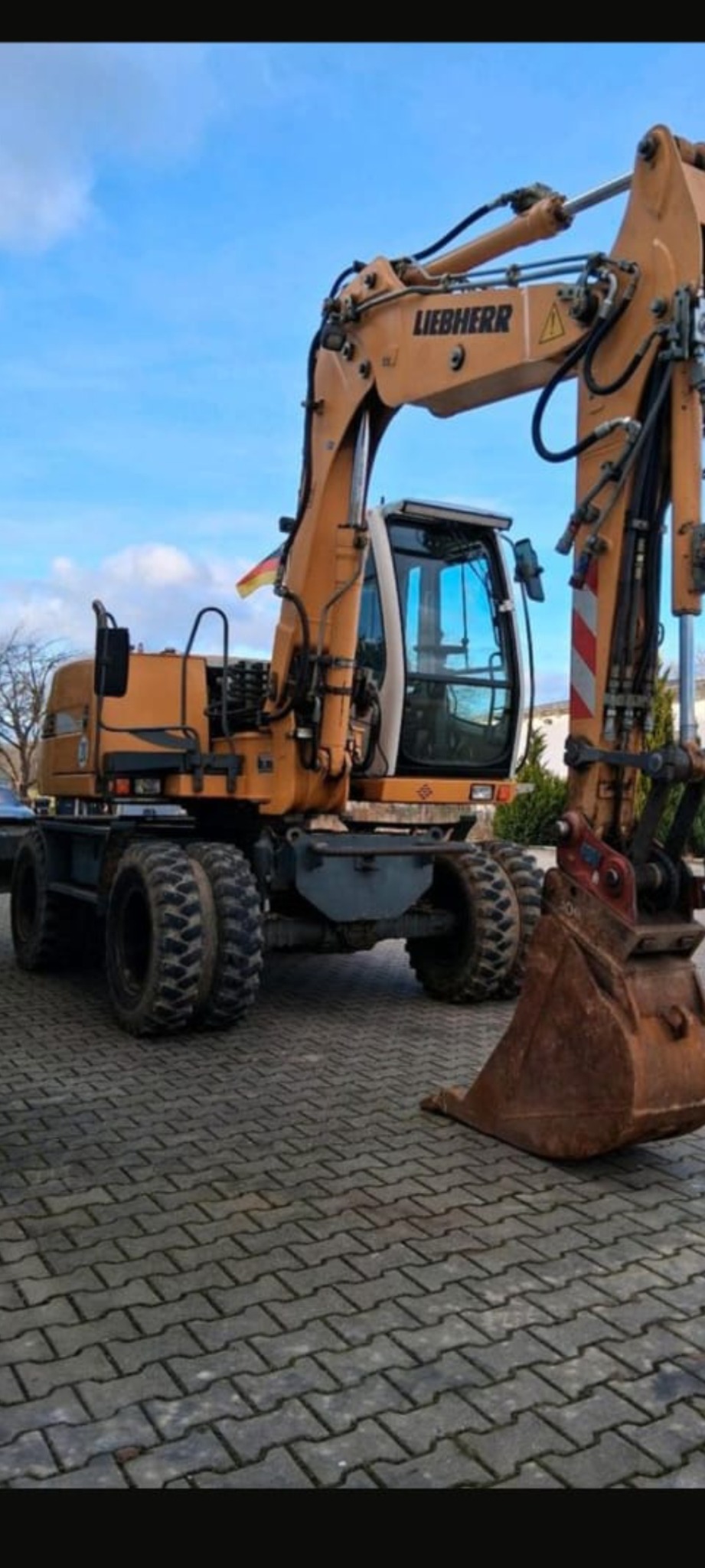 Żółta koparka Liebherr na kostce brukowej, z niemiecką flagą. Widoczne opony, kabina i ramię koparki. Błękitne niebo w tle. Maszyna budowlana w trakcie pracy.