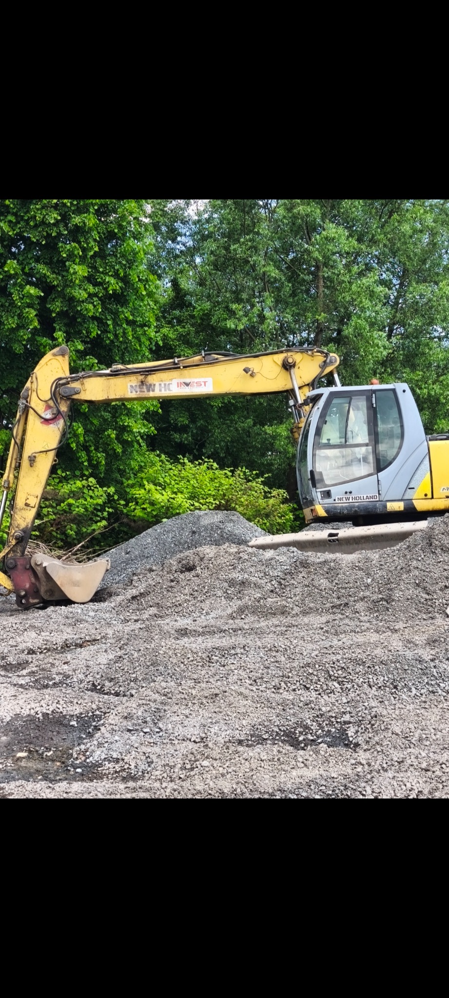Żółto-szara koparka New Holland na tle zieleni, stoi na stercie gruzu. Widoczne logo firmy NEW HC INVEST na ramieniu koparki. Sprzęt budowlany w trakcie pracy.