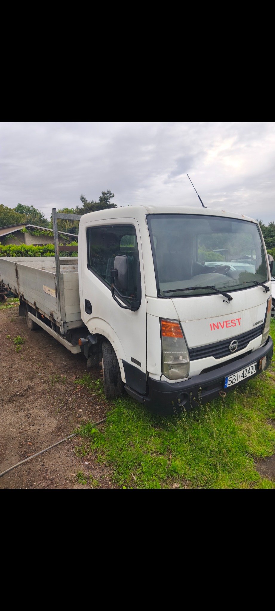 Biała ciężarówka Nissan Cabstar z napisem INVEST na tle zieleni i pochmurnego nieba. Widoczny numer rejestracyjny. Ujęcie z boku, na trawiastym terenie.