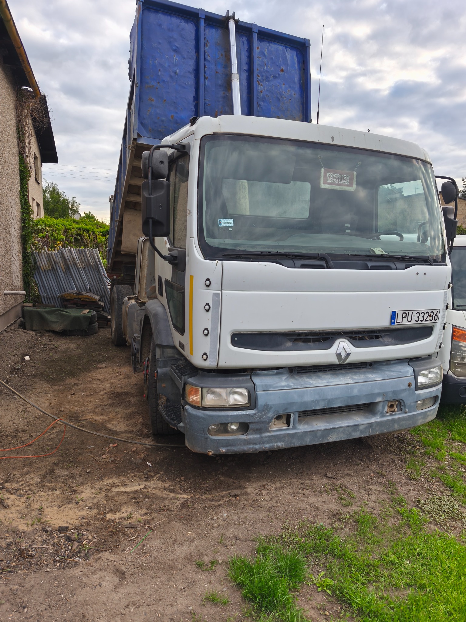 Ciężarówka Renault z podniesioną, niebieską skrzynią ładunkową, zaparkowana na placu budowy. Widoczny numer rejestracyjny i elementy otoczenia.