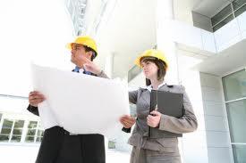 Dwoje inżynierów budowlanych w żółtych kaskach analizuje plan architektoniczny na tle nowoczesnego budynku biurowego z dużymi oknami.