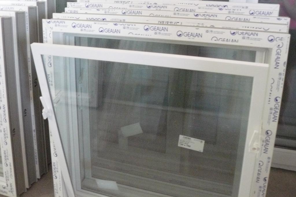 Białe okno PCV marki Gealan, uchylone, z widoczną szybą i naklejką z wymiarami, ustawione obok innych okien w magazynie lub sklepie.