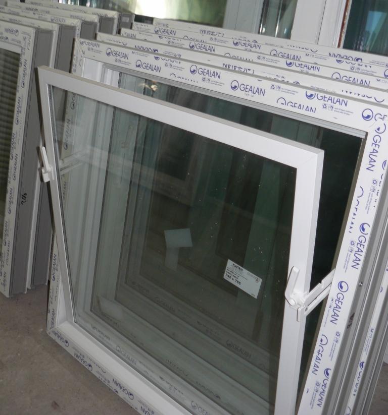 Białe, częściowo otwarte okno PCV z szybą zespoloną, widoczne logo GEALAN na ramie. W tle inne okna, ułożone jedno za drugim. Na szybie naklejka z rozmiarem.