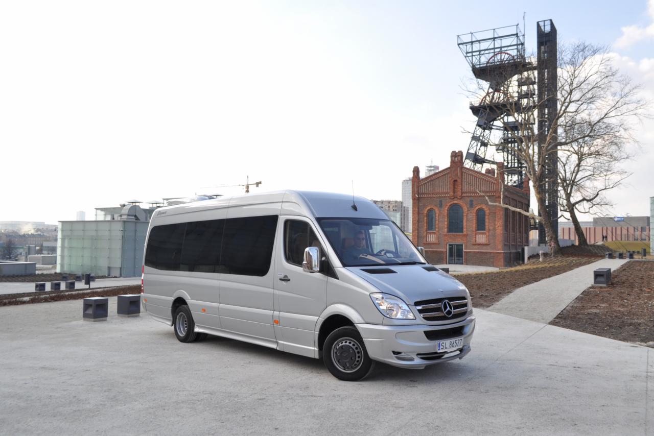 Srebrny bus Mercedes-Benz z przyciemnianymi szybami na tle industrialnego krajobrazu z ceglaną wieżą i konstrukcją stalową.