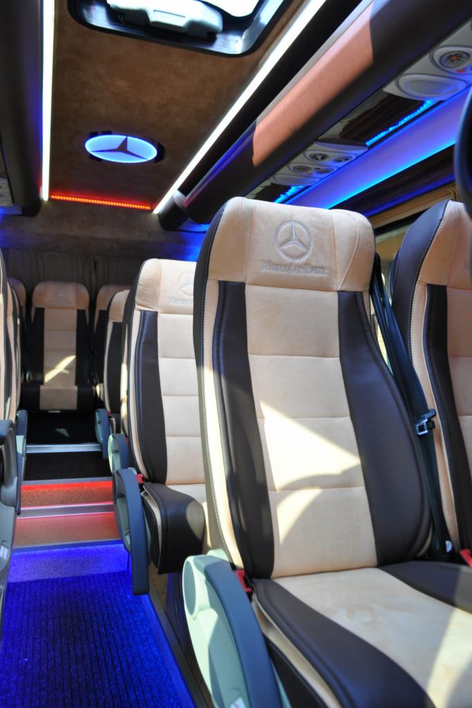 Wnętrze luksusowego busa marki Mercedes-Benz z beżowo-brązowymi fotelami, oświetleniem LED i logiem producenta na zagłówkach. Widok na rząd siedzeń.