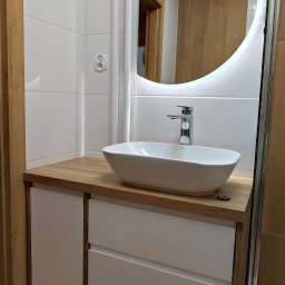 fresno Adam Pietrzak - Nowoczesna łazienka z białą umywalką nablatową, szafką z drewnianym blatem i okrągłym lustrem z podświetleniem LED, widoczny fragment kabiny prysznicowej i narzędzia pod szafką.