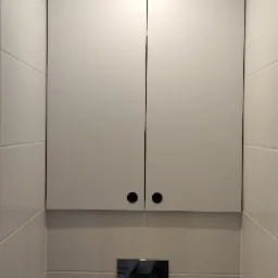 Biała szafka wisząca z czarnymi okrągłymi uchwytami nad spłuczką w toalecie wyłożonej jasnymi, kwadratowymi płytkami. Oświetlenie punktowe nad szafką.