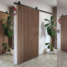 fresno Adam Pietrzak - Drewniana, przesuwna ścianka działowa z pionowych listew, zamontowana na czarnej szynie, częściowo zasłaniająca wejście, z rośliną doniczkową obok, na podłodze z płytek imitujących drewno.