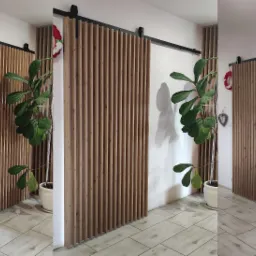 Drewniana, przesuwna ścianka działowa z pionowych listew, zamontowana na czarnej szynie, częściowo zasłaniająca wejście, z rośliną doniczkową obok, na podłodze z płytek imitujących drewno.