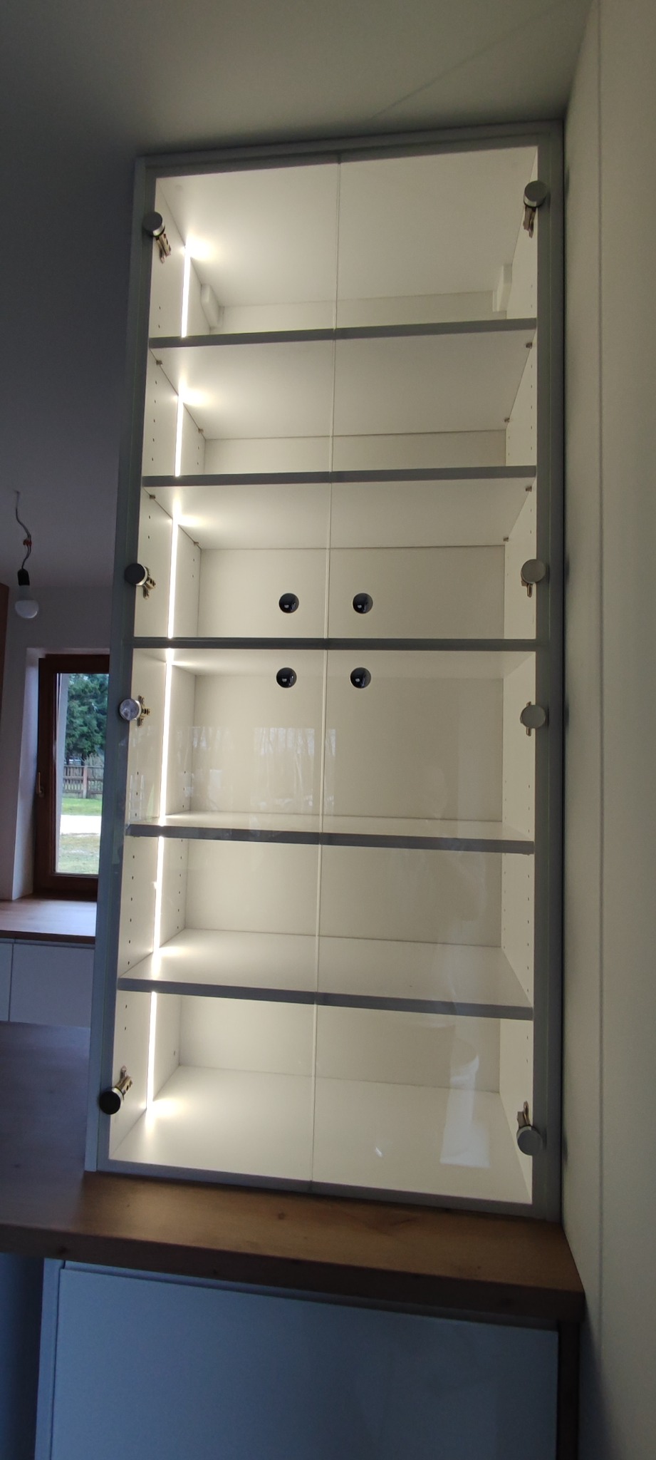 Wysoka, biała szafka kuchenna z szklanymi drzwiami, oświetlona pionowymi listwami LED, zamontowana na drewnianym blacie.