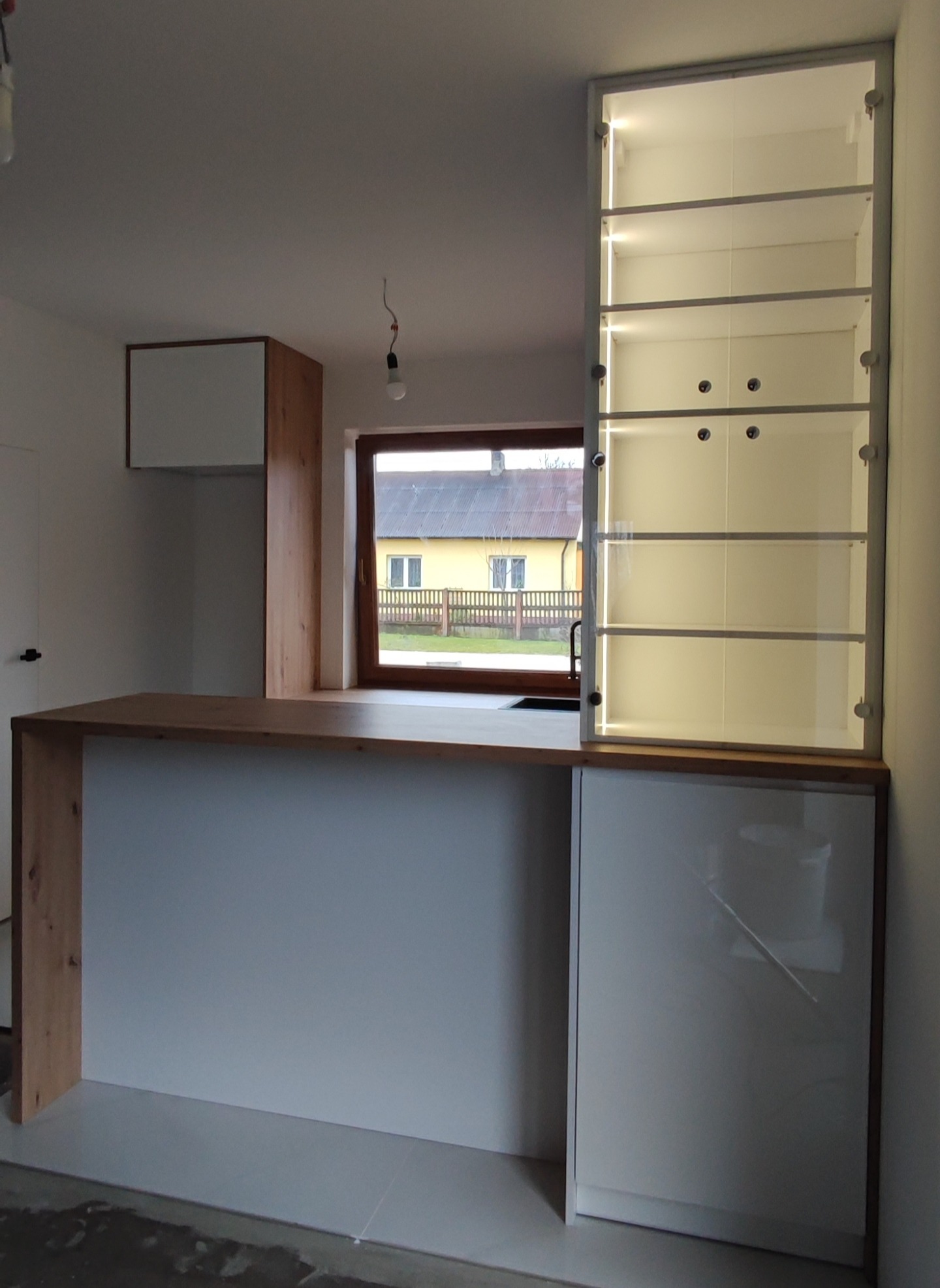 Biała kuchnia na wymiar z drewnianym blatem i oświetloną witryną w Zduńskiej Woli, widok na okno z zabudową kuchenną.