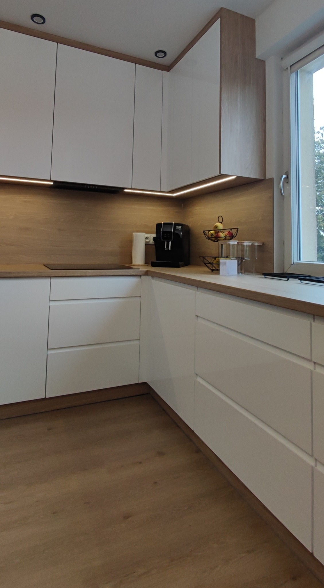 Nowoczesna kuchnia w odcieniach bieli i drewna z podświetleniem LED, ekspres do kawy na blacie, widok na okno z zielenią za szybą.