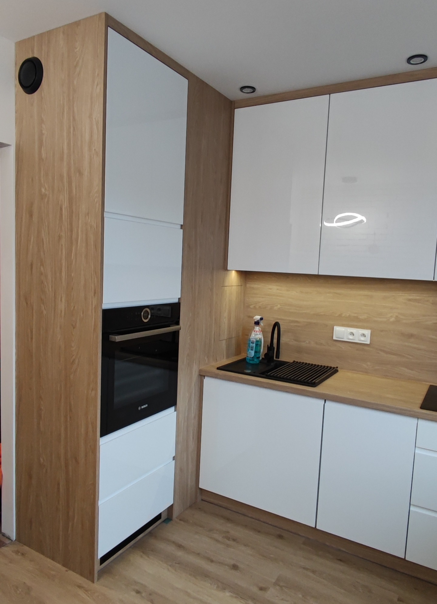 Nowoczesna kuchnia w Zduńskiej Woli z białymi frontami i drewnopodobnym wykończeniem, widoczny piekarnik w zabudowie oraz czarny zlew z ociekaczem.