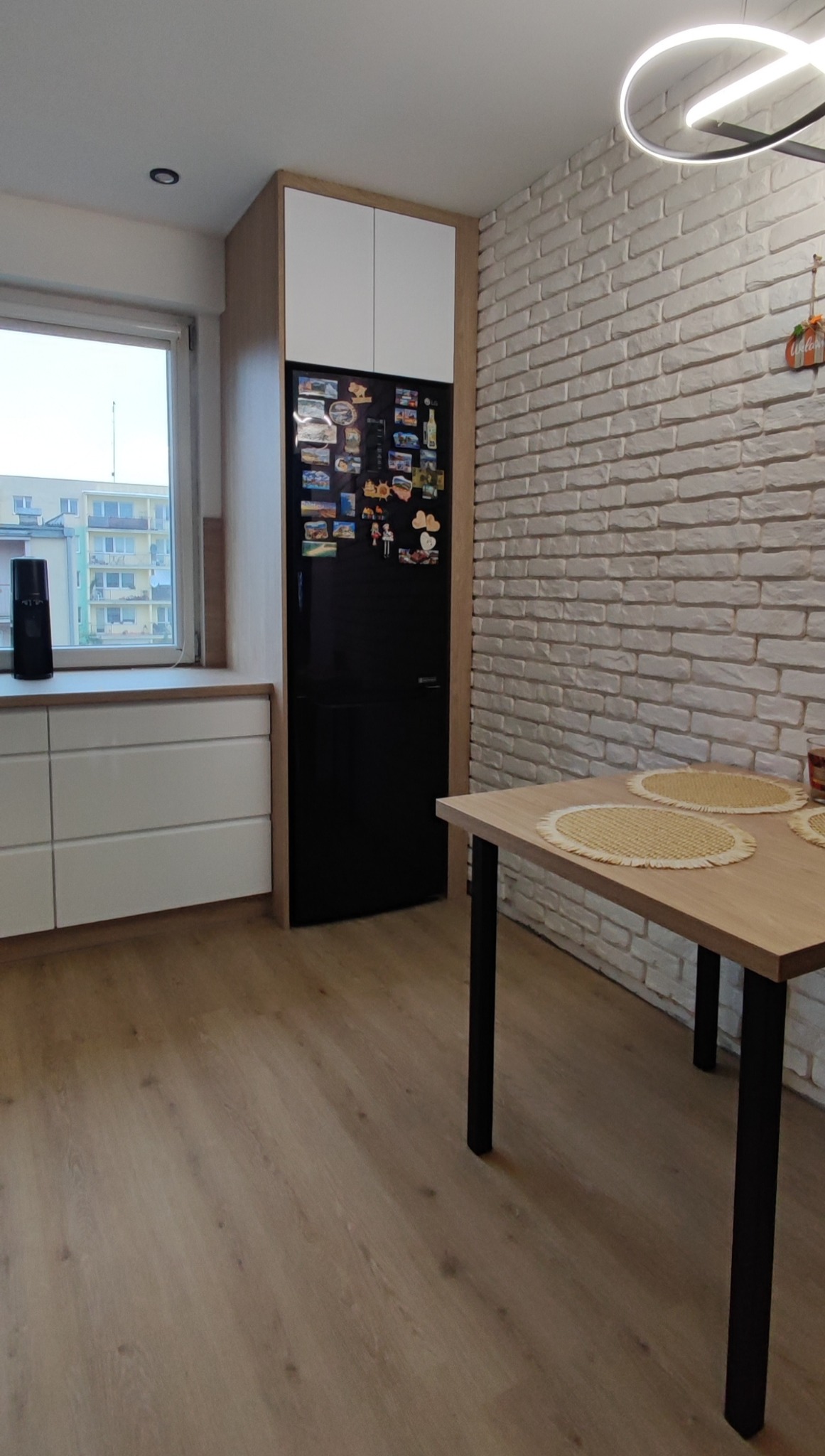 Wnętrze kuchni z czarną lodówką w zabudowie, białymi szafkami, stołem z czarnymi nogami i ścianą z białej cegły dekoracyjnej.