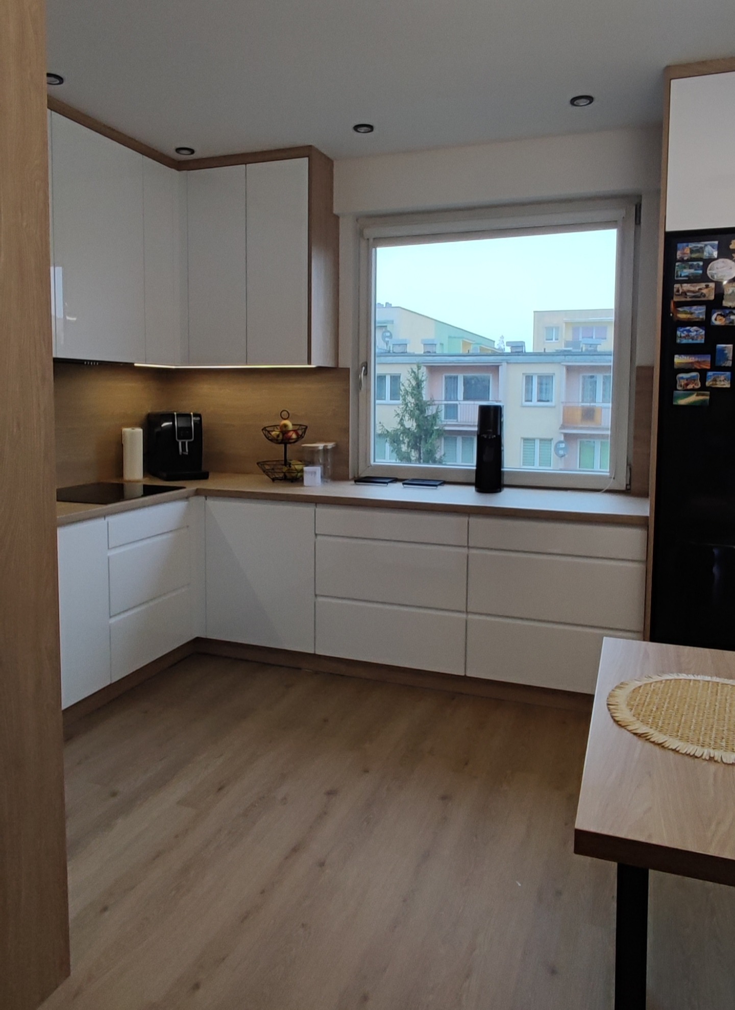 Nowoczesna, jasna kuchnia z białymi szafkami i drewnianym blatem, widok na budynki przez okno, na blacie ekspres do kawy i koszyk z owocami, na lodówce magnesy.