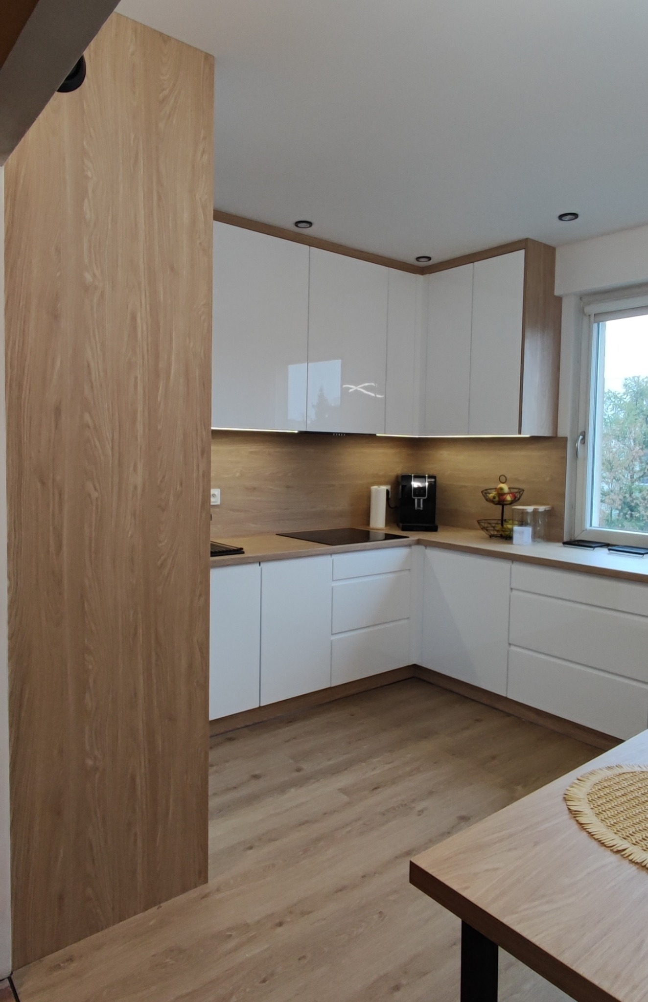 Nowoczesna, jasna kuchnia z białymi szafkami i drewnianymi akcentami, oświetleniem LED pod szafkami wiszącymi, widok na okno i fragment stołu z plecioną podkładką.