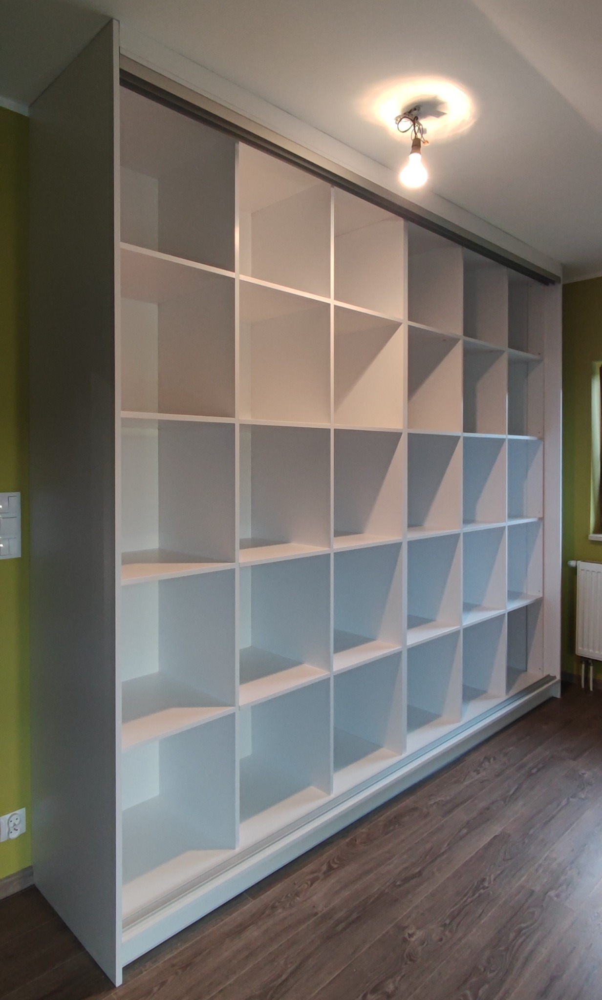 Biała zabudowa meblowa z licznymi półkami w kształcie kwadratów, widoczne przesuwne drzwi, podłoga z paneli imitujących drewno, na suficie żarówka bez klosza.